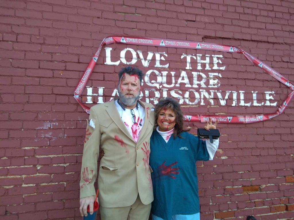 Photo of zombie man in a torn suit and zombie woman wearing scrubs in front of Brick Wall with the words, "Love the Square Harrisonville" on it. 