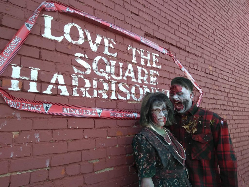 Photo of zombie man in torn plaid shirt and zombie woman in a torn 1980's dress in front of Brick Wall with the words, "Love the Square Harrisonville" on it. 